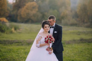 Gelin ve damat bir tarlada duruyorlar, gelinin elinde bir buket kırmızı çiçek var. Çiftler düğünlerini kutlarken sahne romantik ve mutlu.