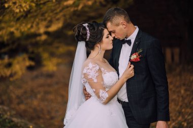 Bir gelin ve damat ormanda sarılıyor. Gelin beyaz bir elbise giyiyor ve damat da takım elbise giyiyor.