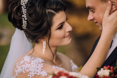 Bir gelin ve damat düğün fotoğrafında birbirlerine sarılıyorlar. Damat takım elbise giyerken gelin beyaz bir elbise ve taç giyiyor. Fotoğraf aşkı yakalar.
