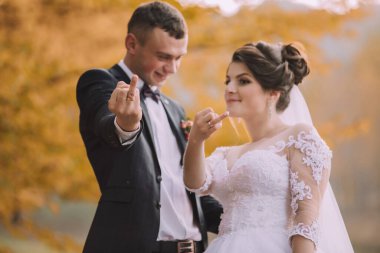 Gelin ve damat, gelinin orta parmağını kaldırdığı bir resim için poz veriyorlar. Adam takım elbise ve kravat takıyor ve gelin beyaz bir elbise giyiyor. Sahne neşeli ve neşeli.