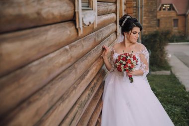 Beyaz elbiseli bir kadın ahşap bir duvarın önünde duruyor. Elinde bir buket kırmızı çiçek tutuyor.