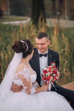 Bir gelin ve damat birlikte oturuyor, gülümsüyor ve el ele tutuşuyorlar. Gelin beyaz bir elbise ve duvak takarken damat takım elbise ve papyon takıyor.