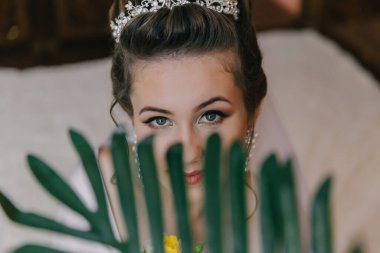 Taclı bir kadın kameraya bakıyor. Beyaz bir elbise giyiyor ve saçları topuz şeklinde.