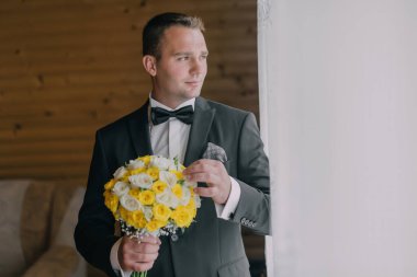 Takım elbiseli bir adam elinde bir buket sarı ve beyaz çiçek tutuyor. Pencereden dışarı bakıyor.