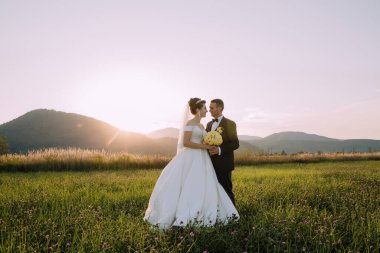 Bir gelin ve damat arka planda dağlarla bir tarlada duruyor. Gelin beyaz bir elbise giyiyor ve damat siyah bir takım elbise giyiyor. El ele tutuşup kameraya gülümsüyorlar.