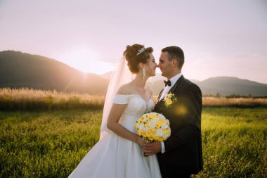 Gelin ve damat bir tarlada duruyorlar, gelin bir buket tutuyor ve damat sarı bir çiçek tutuyor. Güneş arka planda batıyor, sıcak ve romantik bir atmosfer yaratıyor.