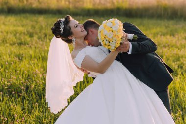 Bir gelin ve damat çimen tarlasında kucaklaşıyor. Gelin beyaz bir elbise giyiyor ve damat bir buket sarı çiçek tutuyor. Sahne romantik ve mutlu.