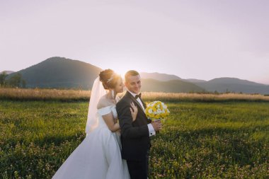 Bir gelin ve damat sarı buketli bir tarlada duruyor. Gelin beyaz bir elbise giyiyor ve damat siyah bir takım elbise giyiyor. Güneş arkalarında batıyor, sahne üzerine sıcak bir parıltı saçıyor.