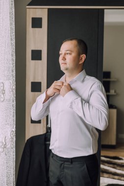 Takım elbiseli ve kravatlı bir adam pencerenin önünde duruyor, yakasını düzeltiyor. Adam takım elbise ve kravat giydiği için resmiyet ve profesyonellik kavramı. Pencere bir arkaplan görevi görür