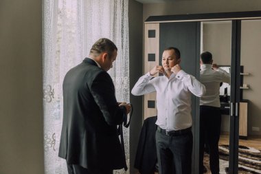 İki adam resmi bir etkinliğe hazırlanıyor. Bir tanesi aynaya bakarken diğeri kravatını düzeltiyor. Sahne ciddi ve odaklı.