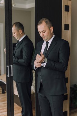 Takım elbiseli bir adam aynanın önünde kravatını düzeltiyor. Formalite ve detaylara dikkat etme kavramı, adamın görünüşünün parlak ve profesyonel olması için zaman ayırması gibi.