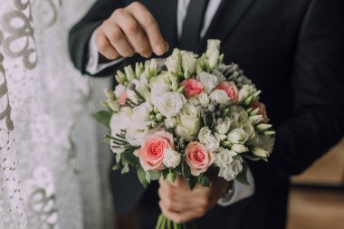 Bir adam elinde bir buket çiçek tutuyor. Buket beyaz ve pembe çiçeklerin bir karışımıdır ve bir vazoya yerleştirilir. Adam takım elbise ve kravat takıyor.