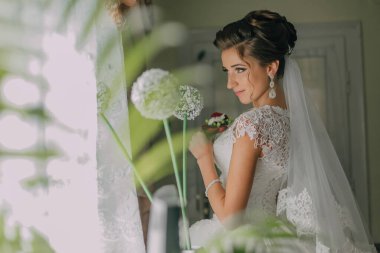 Bir gelin elinde bir buket çiçekle pencerenin önünde duruyor. Beyaz bir elbise ve duvak takıyor.