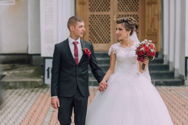 Bir gelin ve damat el ele tutuşup gülümsüyor. Damat takım elbise giyerken gelin beyaz bir elbise ve kırmızı bir kravat takıyor. İkisi de bir buket taşıyor.