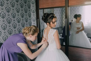 Bir kadın düğününe hazırlanıyor ve nedimesi ona yardım ediyor. Gelin beyaz bir elbise giyiyor ve nedime mor bir elbise giyiyor.