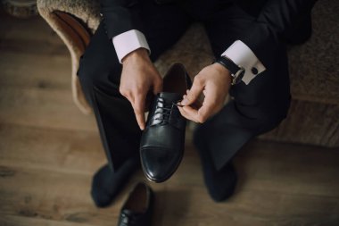 Takım elbiseli bir adam ayakkabılarını giyiyor. Saat takıyor ve elleri ayakkabısında.