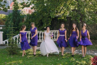 Bir gelin ve nedimeleri parkta bir patikada yürüyorlar. Gelin beyaz bir elbise giyiyor ve nedimeler mor elbiseler giyiyor. Gelin bir buket tutuyor.