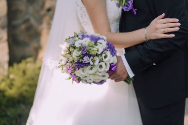 Bir gelin ve damat bir buket mor ve beyaz çiçek tutarken birbirlerini kucaklıyorlar. Çiçekler bir vazoda sergileniyor ve çift düğün kıyafeti giyiyor. Sahne aşktır.