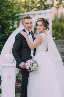 Gelin ve damat beyaz bir merdivende fotoğraf çektirmek için poz veriyorlar. Gelin beyaz bir elbise giyiyor ve damat siyah bir takım elbise giyiyor. Gelin elinde bir buket çiçek tutuyor.