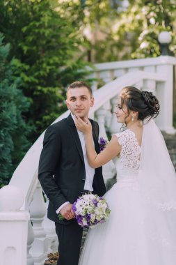 Gelin ve damat köprüde duruyorlar, gelin elinde bir buket mor çiçek tutuyor.