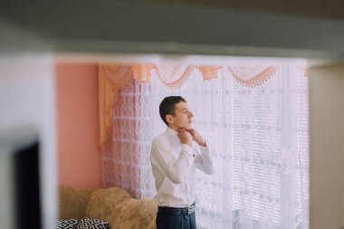 Beyaz gömlekli bir adam giyinmeye hazırlanıyor. O bir pencerenin önünde duruyor ve odanın pembe ve turuncu renkli bir düzeni var.