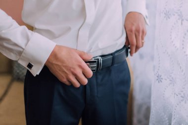 Bir adam beyaz gömlek ve mavi pantolon giyiyor. O, kemerini ayarlıyor. Formalite kavramı ve detaylara dikkat