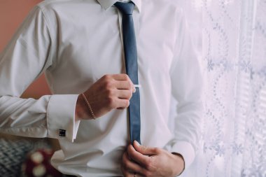 Bir adam işe hazırlanıyor, mavi kravat takıyor. Beyaz bir gömlek ve altın bir bilezik takıyor.