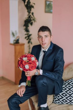 Elinde bir buket kırmızı gül tutan takım elbiseli bir adam. Kanepede oturuyor.