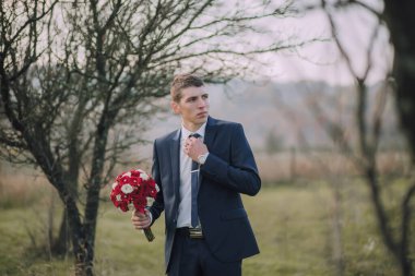 Takım elbise giymiş ve kravat takmış bir adam elinde bir buket kırmızı çiçekle bir tarlada duruyor. Sahne sakin ve huzurlu, adam uzaklığa bakıyor.