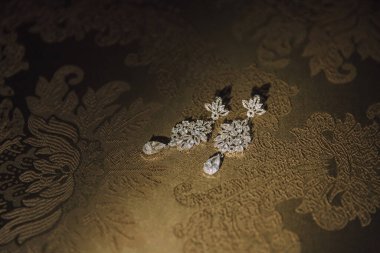 Çiçek desenli bir çift küpe bir masanın üzerinde oturuyor. Küpeler elmastan yapılmış ve altın çerçeveye yerleştirilmiş. Masa desenli bir kumaşla kaplı.