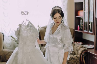 Bir gelin gelinliğin önünde duruyor ve büyük günü için hazırlanıyor. Elbise beyaz ve üzerinde bir taç var. Sahne bir oturma odasında geçiyor, yanında bir kanepe ve sandalye var.