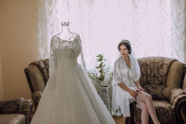 Bir gelin, bir kanepede gelinliğin önünde oturuyor. Elbise bir mankene asılı.