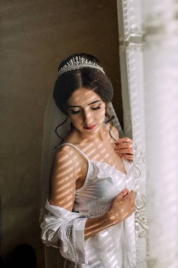 Beyaz elbiseli bir kadın pencerenin önünde duruyor, kolları çapraz ve kafasında bir peçeyle. Şık ve sofistike bir kadın düğün kıyafeti giydiği için.