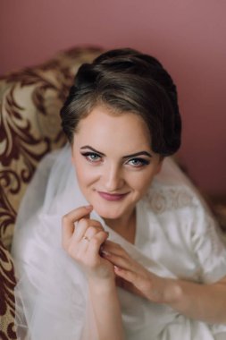 Beyaz bornozlu bir kadın sandalyede oturuyor. Gülümsüyor ve eli yüzünde.