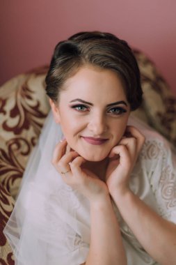 Bir kadın elleri yüzü üzerinde bir sandalyede oturuyor. Gülümsüyor ve saçları topuz gibi.