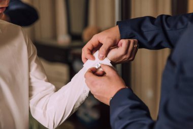 İki adam giyiniyor, biri diğerine kelepçe takıyor. Kelepçe takan adam beyaz bir gömlek giyiyor ve diğeri mavi bir takım giyiyor.