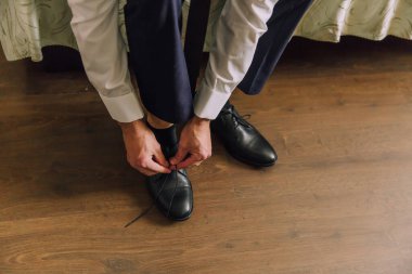 Bir adam ayakkabılarını giyiyor. Ayakkabılar siyah ve adam mavi bir takım elbise giyiyor.