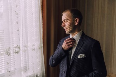 Takım elbiseli bir adam kravatını bağlamaya hazırlanıyor. O bir pencerenin önünde duruyor ve güneş ışığı onun üzerinde parlıyor. Formalite ve profesyonellik kavramı