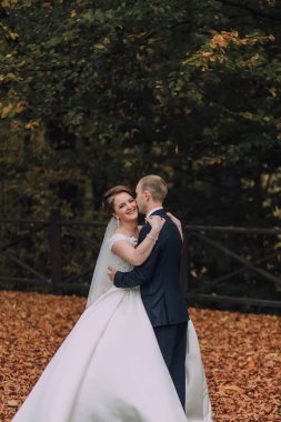 Bir gelin ve damat ormanda sarılıyor. Gelin beyaz bir elbise giyiyor ve damat mavi bir takım giyiyor.