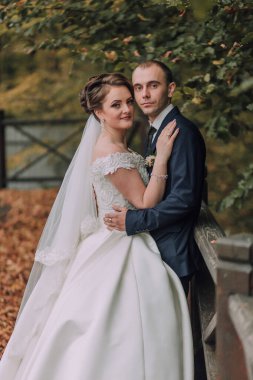 Bir gelin ve damat bir ağacın önünde fotoğraf için poz veriyorlar. Gelin beyaz bir elbise giyiyor ve damat mavi bir takım elbise giyiyor. Sahne mutlu ve romantik.