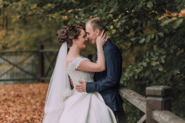 Bir gelin ve damat köprüde birbirlerine sarılıyorlar. Gelin beyaz bir elbise giyiyor ve damat mavi bir takım elbise giyiyor. Sahne mutlu ve romantik.