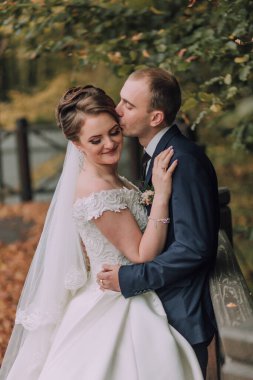 Bir gelin ve damat köprüde öpüşüyorlar. Gelin beyaz bir elbise giyiyor ve damat mavi bir takım giyiyor.