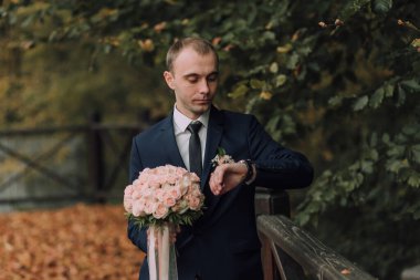 Takım elbiseli ve kravatlı bir adam saat takıyor ve elinde bir buket pembe çiçek tutuyor. Saati kontrol ediyor, muhtemelen bir etkinliğe ya da toplantıya hazırlanıyor.