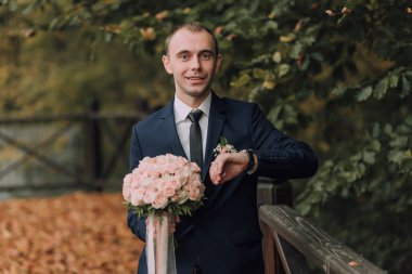 Takım elbiseli ve kravatlı bir adam bir buket pembe çiçek tutuyor. Saat takıyor ve gülümsüyor.