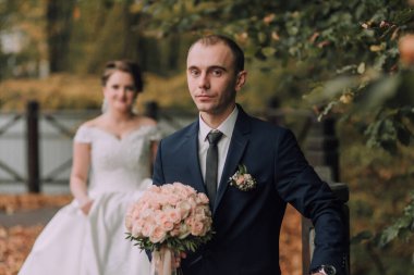 Bir adam ve bir kadın parkta duruyorlar. Adam takım elbise ve kravat takıyor ve kadın beyaz bir elbise giyiyor. Adam elinde bir buket çiçek tutuyor ve kadın ona bakıyor.