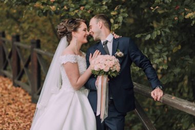 Bir gelin ve damat köprüde el ele tutuşup gülümsüyor. Gelin beyaz bir elbise giyiyor ve damat mavi bir takım elbise giyiyor. İkisinin de elinde çiçekler var.