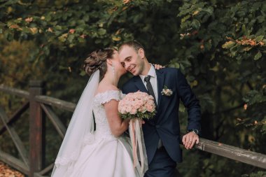 Bir gelin ve damat köprüde birbirlerine sarılıp öpüşüyorlar. Gelin beyaz bir elbise giyiyor ve damat mavi bir takım giyiyor.