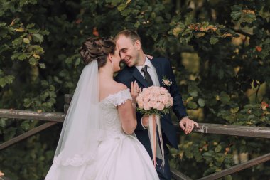 Bir gelin ve damat köprüde kameraya gülümsüyor. Gelin beyaz bir elbise giyiyor ve damat mavi bir takım elbise giyiyor. Bir buket çiçek ve çiçek tutuyorlar.