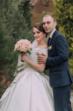 Bir gelin ve damat bir ağacın önünde fotoğraf için poz veriyorlar. Gelin bir buket çiçek tutuyor ve damat takım elbise giyiyor. Sahne mutlu ve kutlama dolu.