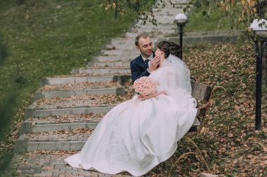 Bir gelin ve damat parkta bir bankta oturuyorlar. Gelin elinde bir buket çiçek tutuyor.